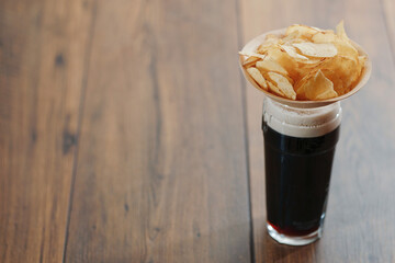 Dark ale in a glass with foam and a wooden small cup with potato chips on a wooden table. Selective focus. Traditional beer and snack