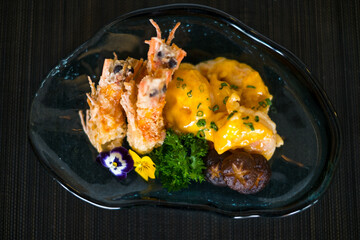 Two shrimps in batter with sauce and mushrooms on the blue designed plate. Deep fried prawns decorated with greenery and flower. Isolated on black