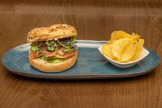 Bread Bagel Traditionally Made From Wheat Flour, Stuffed With Smoked Norwegian Salmon With Avocado Slices And Assorted Lettuce Shoots Garnished With Potato Chips.