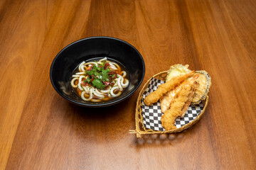 Black bowl of udon noodles with vegetables and tempura plate of prawns and vegetables on wicker basket on wooden table