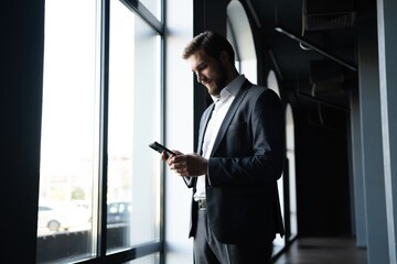 Confident young businessman using smartphone while standing near the window an office building.