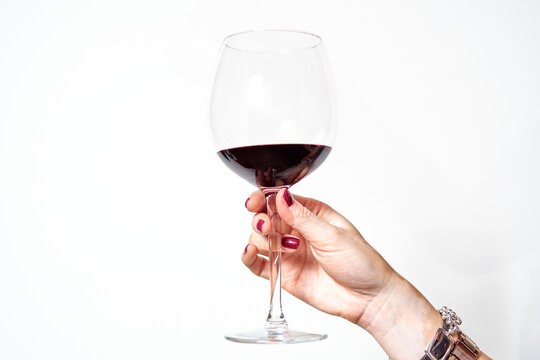 hand of a young woman with vitiligo and red painted nails toasting with a glass of red wine on white background.