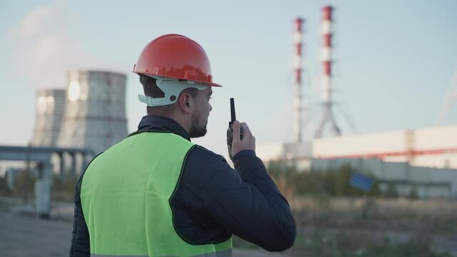Male caucasian engineer in orange helmet and reflective vest talking via transmitter with security workers. Energy power station area with bright sky. Concept of industry, success