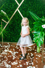  Little cute girl with blond hair in a white dress  and white flowers, lilies and orchids on a  background with a green tropical plants