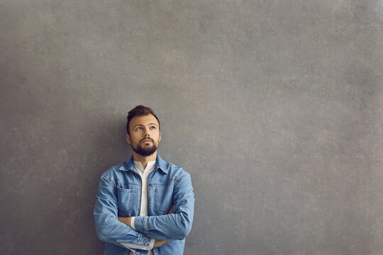 Handsome caucasian man with thinking expression standing on gray concrete background. Man in a denim jacket stands with folded arms and looks up thoughtfully. Advertising concept. Place for text.