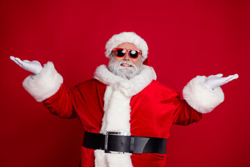 Photo of cheerful man two hands demonstrate empty space variants wear santa hat costume isolated red color background