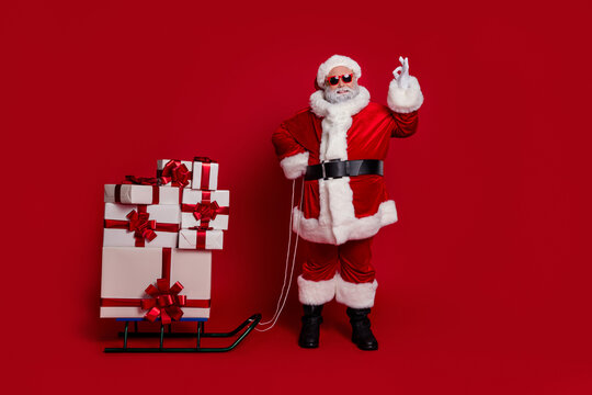Full length photo of funky santa show okey bring gifts by sled wear cap glasses coat pants shoes isolated on red color background