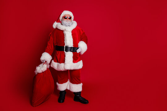 Full length photo of cool santa hold bag with presents wear hat eyewear coat pants boots isolated on red background
