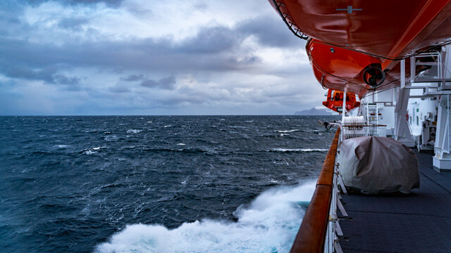 Schiff im Sturm mit hohen Wellen, Gischt und Blick auf ein Rettungsboot. Schifffahrt mit einem Linienschiff der norwegischen K&uuml;ste entlang. Nordatlantik mit Sund und Fjord. Rotes Rettungsboot.