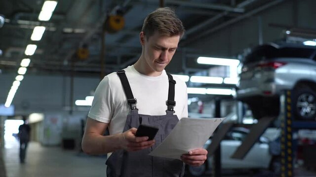 Portrait of serious handsome mechanic male in blue uniform dialing customer number for call. Auto technician text messaging on cell phone in workshop holding report. Tracking shot in slow motion.