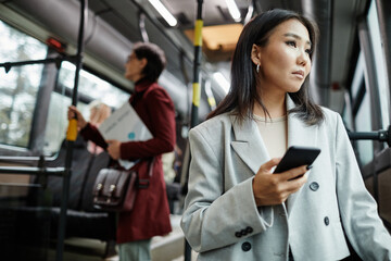 Portrait of elegant Asian woman looking at window on bus while traveling by public transport and holding smartphone, copy space