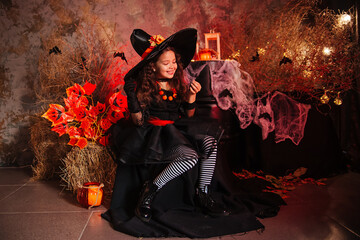 A little girl in a witch costume on a gloomy background holds a black spider in her hands. Getting...