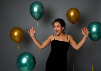 Cheerful young Latin woman in black evening dress rejoices, smiles toothy smile looking at camera against a festive ornate gray wall background with beautiful shiny gold and green metallic balloons