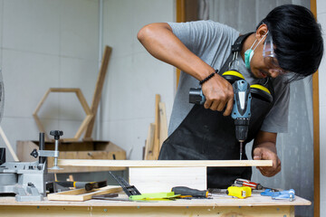 A young male carpenter working on his workshop table using a drill machine and wearing safety equipment