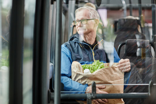 Senior man looking at window in bus while traveling by public transport in city and holding paper bag with groceries, copy space