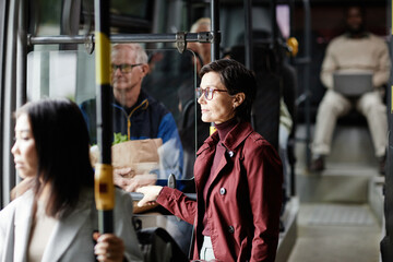 Side view portrait of adult woman holding onto railing in bus while traveling by public transport...