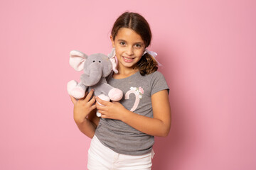 Fototapeta na wymiar childhood, toys and people concept - cute little girl hugging elephant isolated over pink background.