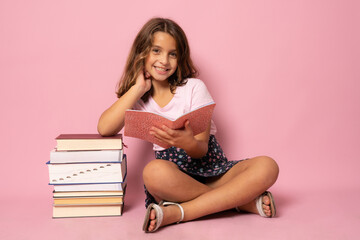 Pretty little girl sit on floor and reading book. Isolated on pink background
