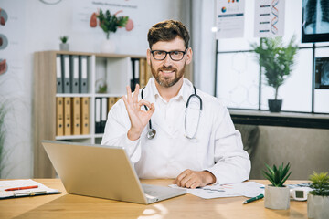 Caucasian male doctor showing approval okay gesture and smiling on camera while sitting at modern office with laptop on table. Modern gadget for medical workers Positive human emotions.