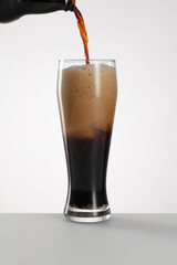 Glass of fresh and cold dark beer on a gray white light background. Stream pouring dark beer from the bottle into a beer glass. Jet of the dark porter pours and whips the thick foam. Selective focus