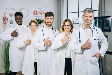 Portrait of happy five multiracial doctors in medical uniform standing at office room, showing their thumbs up and smiling on camera. Concept of people, medicine and health care.