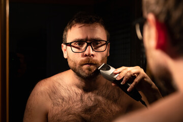 A young man shaving and trimming his beard with an electric razor, in the bathroom at home, early in the morning, to be groomed before going out