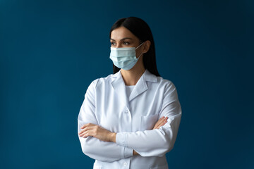 Portrait of attractive serious female doctor wearing protective mask, white lab coat and stethoscope and posing arms crossed isolated blue color background