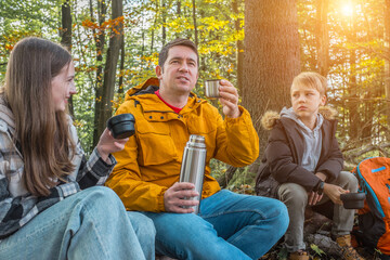 Hikers Father, daughter and son resting in nature forest, drinking hot tea thermos in sunny autumn...