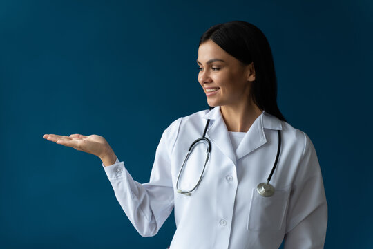 Portrait photo of attractive friendly family doc lady wearing white lab coat and stethoscope smiling reliable and showing with her palm at the empty area isolated blue color background