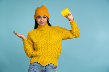 Confused lovely young woman holding empty inverted mug over blue background. Female person feeling disappointed of the empty cup