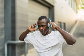 Chico negro atlético con cascos inalámbricos celestes escuchando música, sonriendo y bailando por la calle