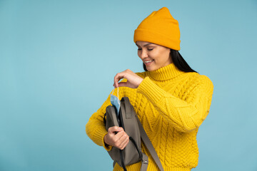 Time for protective mask. Smiling brunette woman wearing yellow knitted sweater and hat taking off from her backpack her protective mask while posing in the studio. Pandemic concept