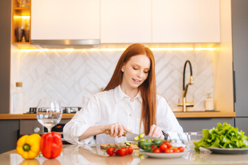 Portrait of attractive young redhead woman cutting fresh cucumber cooking food salad sitting at table in modern kitchen room. Happy female cooking vegetarian dieting salad full of vitamins.