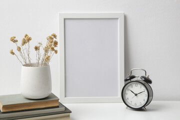 Horizontal white frame mockup on table with books, alarm clock and ceramic vase with yellow dry Everlasting flowers. White wall background.