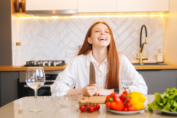 Obraz na płótnie Canvas Portrait of laughing attractive redhead young woman sitting at wooden table with fresh vegetables in light kitchen room with modern interior, holding big knife, closed eyes.