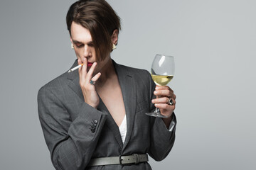 young transgender man in blazer holding glass of wine and smoking isolated on grey