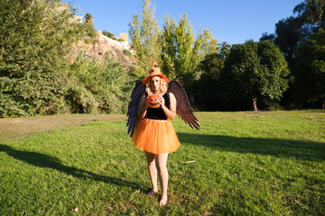 Obraz premium adult woman with blonde hair and a bit chubby in the park enjoying the halloween party. The woman is wearing black lingerie with black wings, an orange witch's hat and a pumpkin to collect candy. 