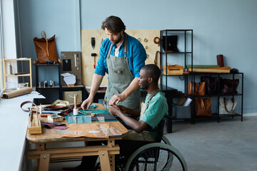 Side view portrait of young African-American man in wheelchair learning leathermaking craft in leatherworkers workshop, copy space