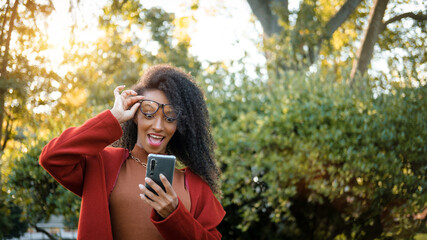 Expressive stylish afro hairstyle black woman surprised while looking at her smartphone outside in autumn.