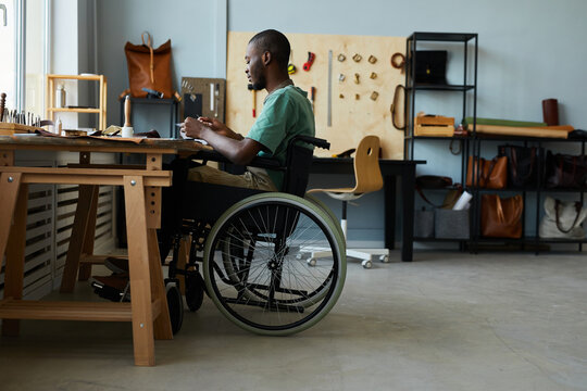 Full length side view portrait of young African-American man in wheelchair making handmade bag in leatherworkers workshop, copy space