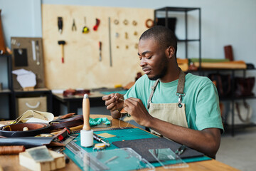 Portrait of young African-American artisan working with leather in leatherworkers workshop, copy...