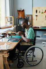 Vertical full length portrait of young African-American man in wheelchair making handmade bag in...