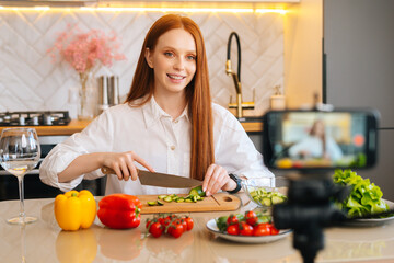 Medium shot portrait of smiling attractive redhead woman vlogger shooting video food blog about cooking on camera of mobile phone, sitting at kitchen with modern light interior, selective focus.