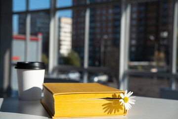 Closed yellow book with a bookmark with a chamomile flower and aromatic coffee in a disposable...