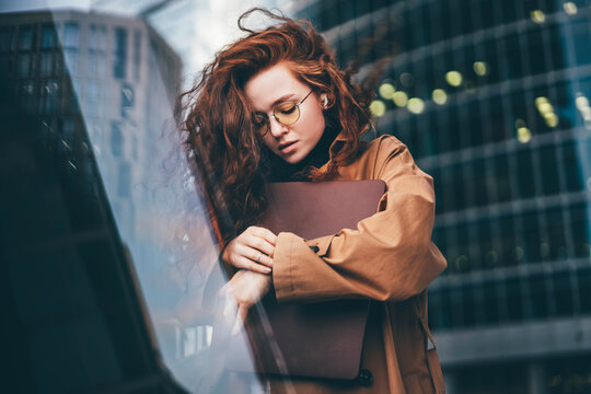Office worker young woman wearing fashion clothes waits for business meeting looking at clock and holds laptop in arms standing against financial center