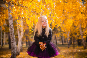 Little cute blonde girl dressed as a witch throws leaves in the park in autumn and smiles. Decorations for All Saints Day. Halloween costume.