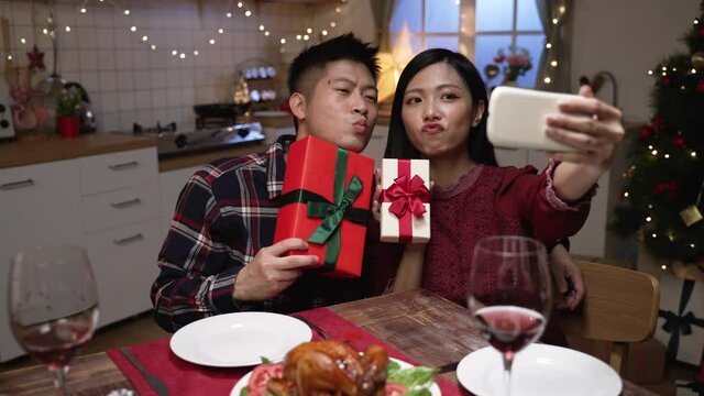cheerful asian couple making funny faces while taking selfie photos with their Christmas presents using mobile phone at xmas eve dinner in dining room