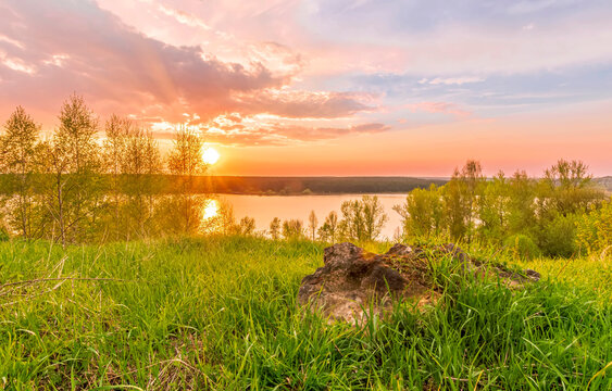 Scenic view at beautiful sunset on a shiny lake with old rough stone on the foreground, green grass, birch trees, golden sun rays, calm water ,nice cloudy sky on a background, spring landscape © Yaroslav