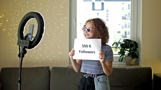 Cute girl blogger shows a poster with 500 thousand subscribers to the camera. The teenager demonstrates his achievements. Blow a kiss to subscribers.
