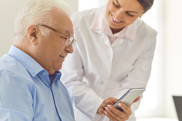 Doctor teaching senior patient to use healthcare app for setting medication reminder or tracking health indicators. Smiling nurse in assisted living facility showing photo of grandchildren to old man
